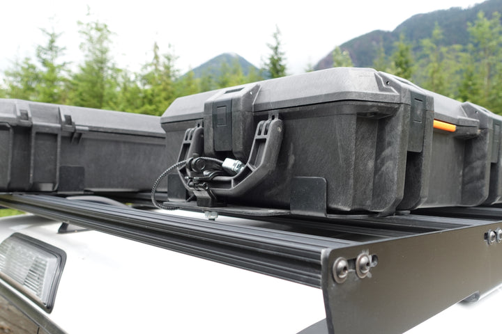 BPF Universal Pelican Vault V700 Case Mounting brackets securely holding a black Pelican Vault V700 case on a vehicle roof rack with a mountainous forest background.