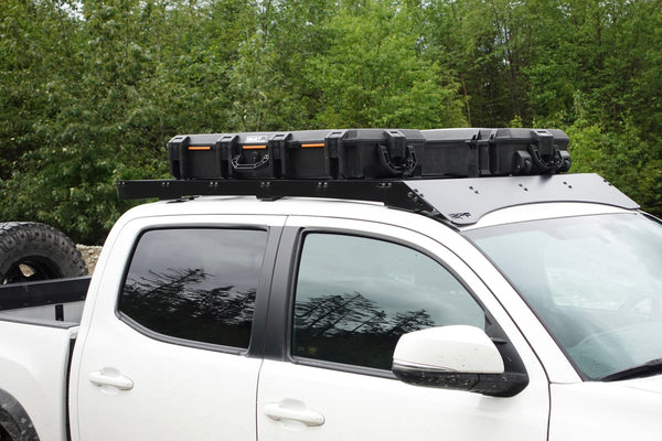 White pickup truck with tinted windows featuring a black roof rack carrying a large rugged storage case, parked outdoors with green trees in the background.