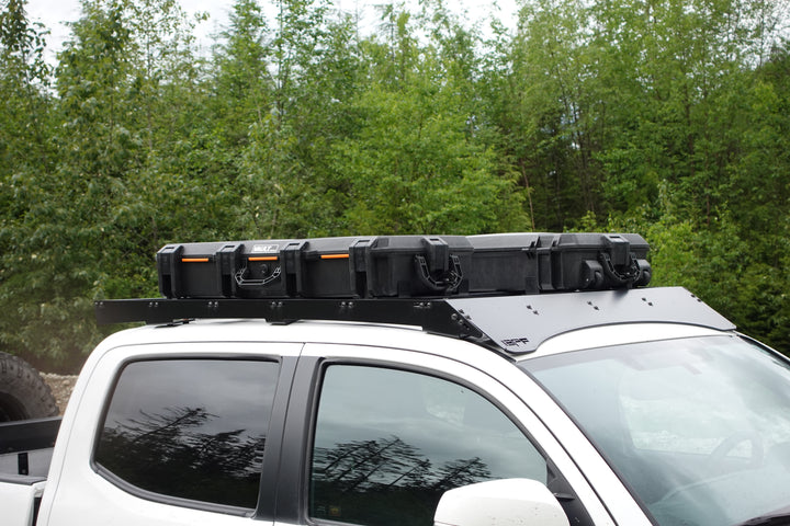 BPF Universal Pelican Vault V800 and V730 Case Mounting brackets securing two black protective cases on a vehicle roof rack against a forest background.