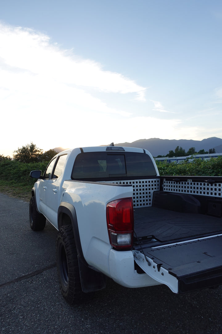 Rear view of a white truck with open tailgate featuring installed BPF 05-23 Tacoma bed Molle panels in a rugged outdoor setting at sunset with mountains in the background