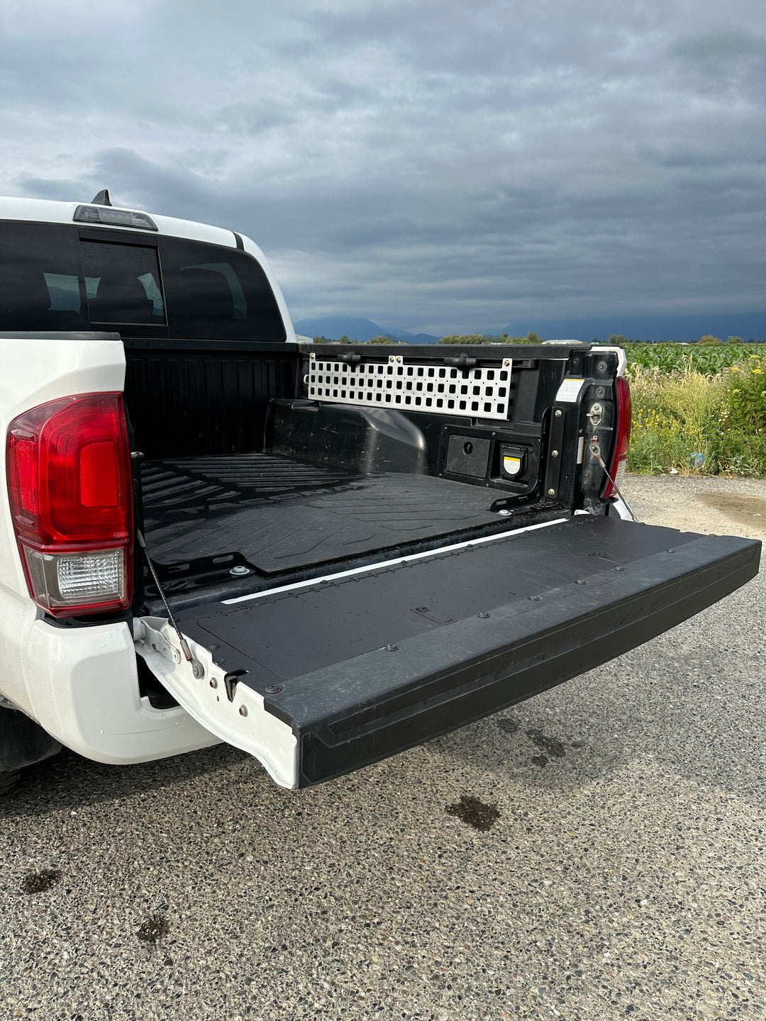 Open truck bed of a white pickup truck showing installed BPF 05-23 Tacoma bed Molle panels on the sidewall, featuring a black protective lining and a white molle panel grid under a cloudy sky.