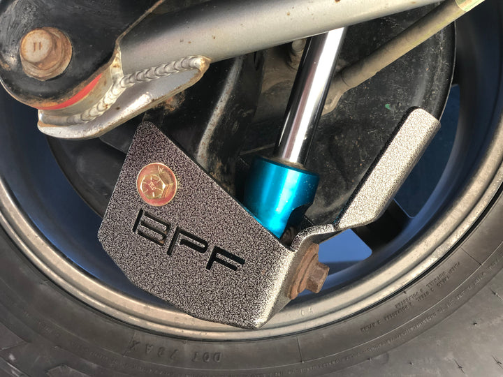 Close-up of a textured metal BPF 4runner 04-24/FJ 07-14 Shock Guards protecting a shock absorber on a vehicle wheel assembly.