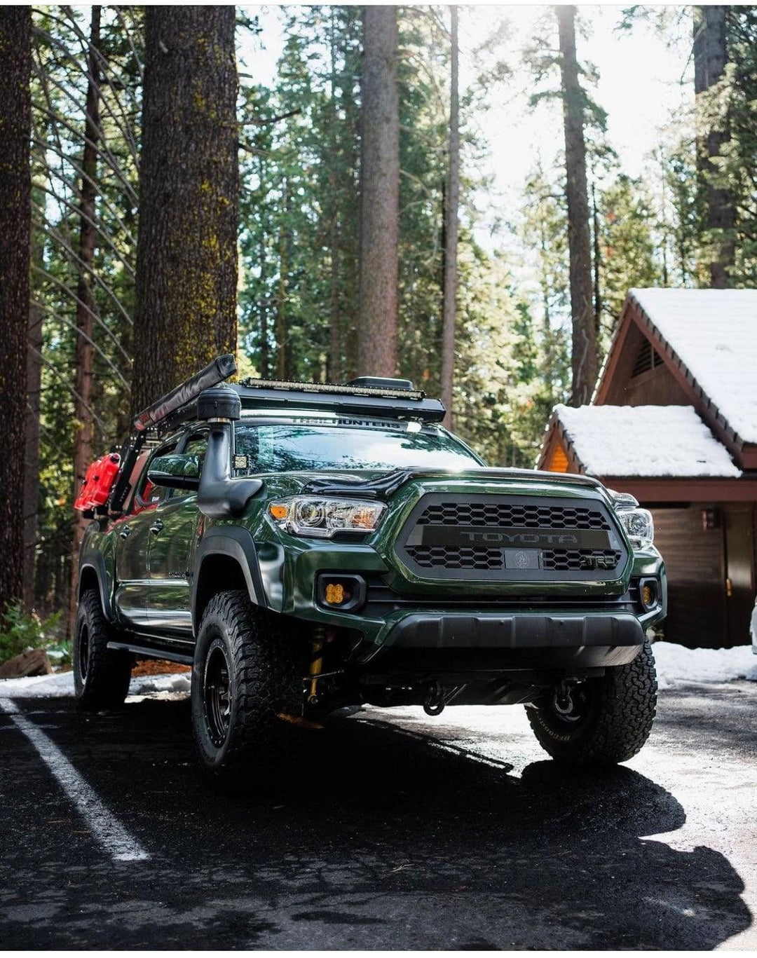 Green Toyota Tacoma equipped with a BPF 2016-2023 Toyota Tacoma Grill, off-road tires, roof rack, and accessories parked in a forested snowy cabin area.