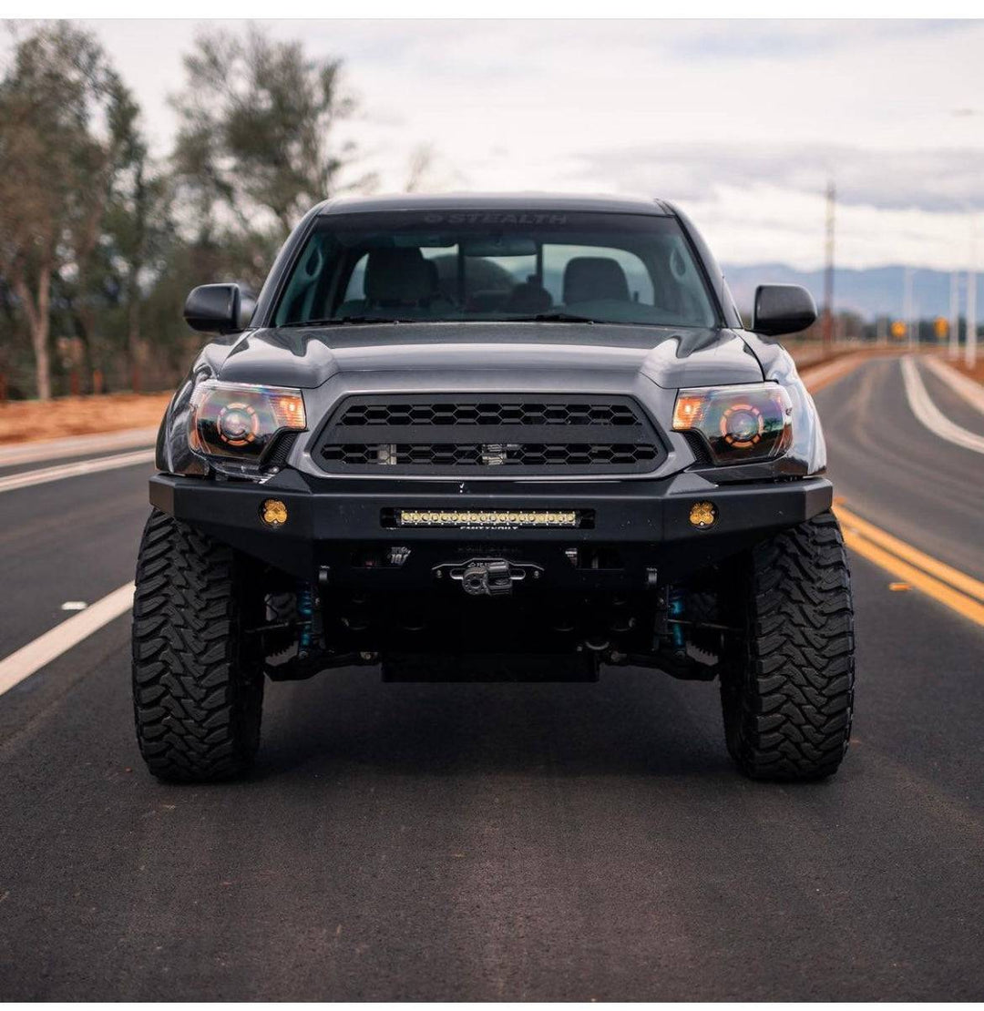 Black Toyota Tacoma truck front view parked on a road featuring a rugged aftermarket BPF 2012-2015 Toyota Tacoma Grill with aggressive off-road tires and a heavy-duty bumper with integrated light bar.